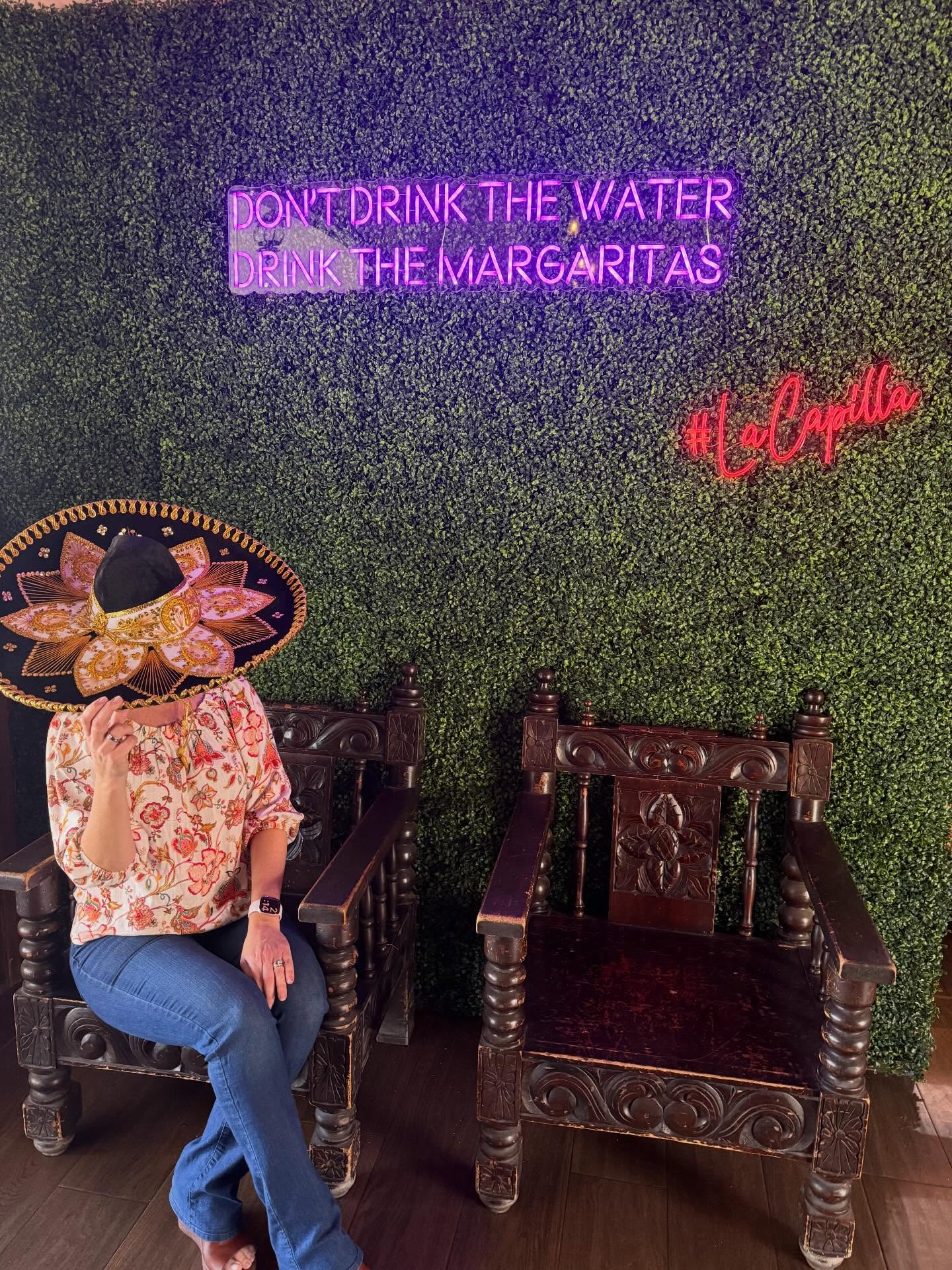 A guest in a sombrero sits beside carved wooden chairs beneath neon signs reading "DON'T DRINK THE WATER, DRINK THE MARGARITAS" and "#LaCapilla."