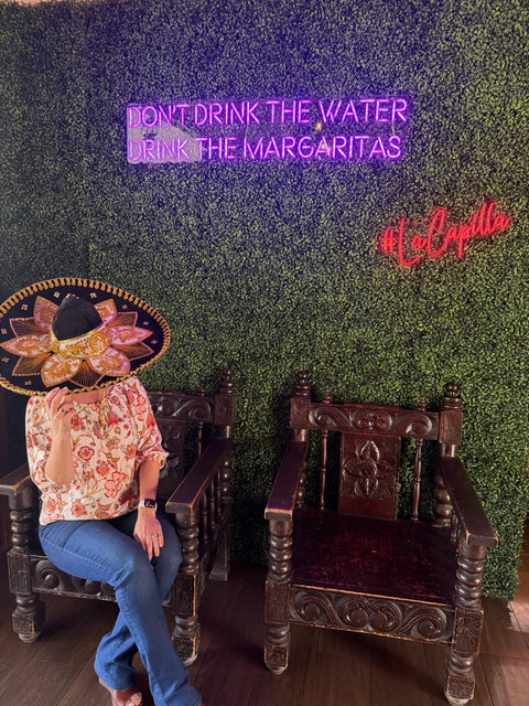 A guest in a sombrero sits beside carved wooden chairs beneath neon signs reading "DON'T DRINK THE WATER, DRINK THE MARGARITAS" and "#LaCapilla."
