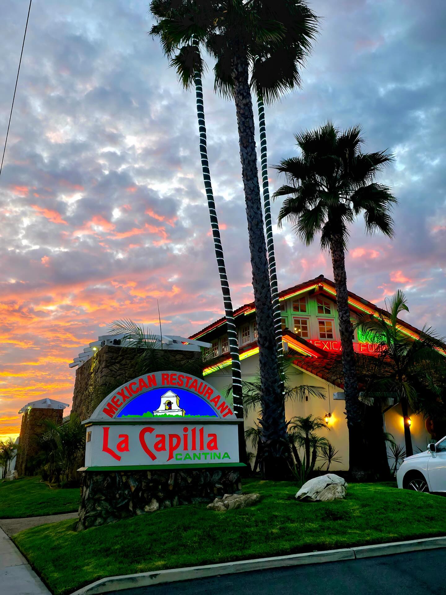 La Capilla restaurant at sunset, with palm trees and an illuminated sign reading "Mexican Restaurant, La Capilla Cantina."