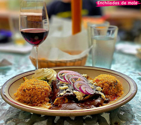 Enchiladas in dark mole sauce topped with red onion and crumbled cheese, served with rice and sour cream. The image reads "Enchiladas de mole."