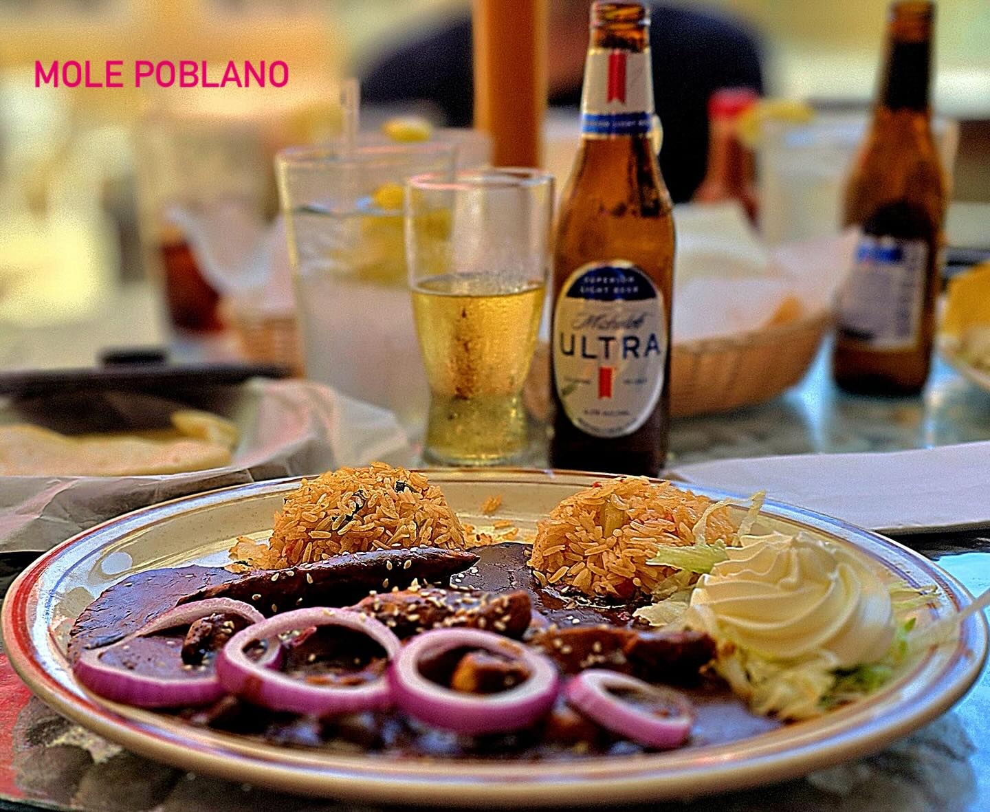 Chicken in dark mole sauce topped with red onion, served with rice, shredded lettuce and sour cream. The image reads "Mole poblano."