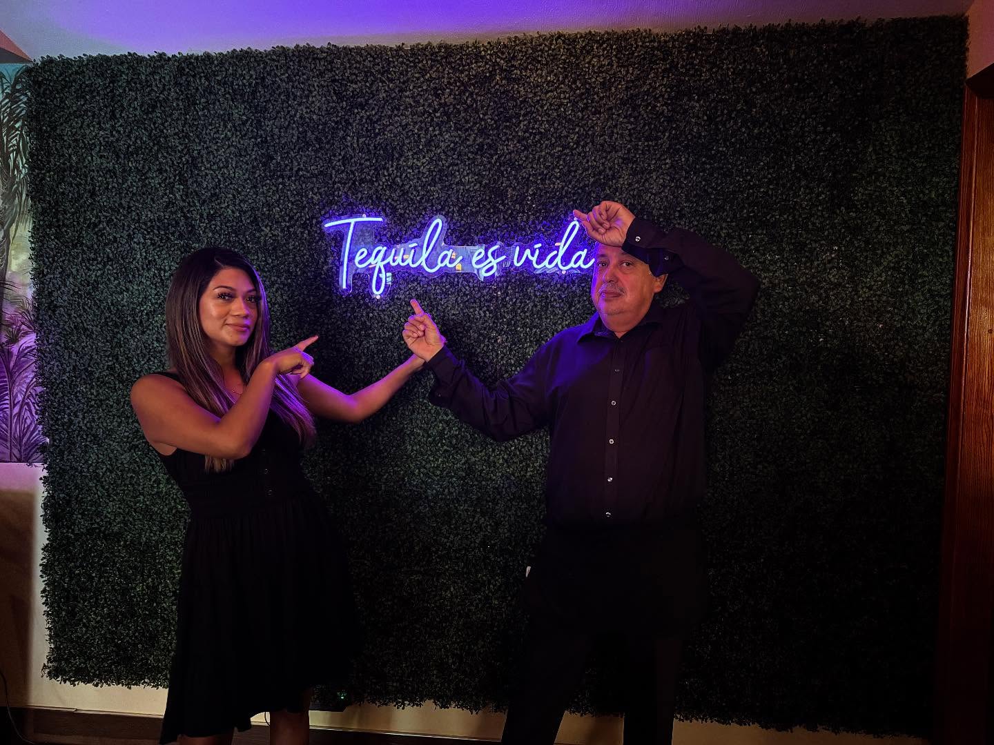 Two guests pose and point toward a purple neon sign reading "Tequila es vida" on a greenery wall.