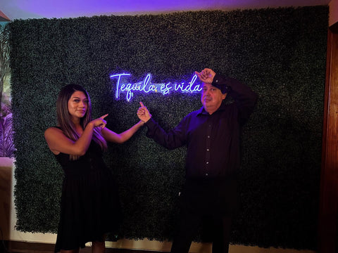 Two guests pose and point toward a purple neon sign reading "Tequila es vida" on a greenery wall.