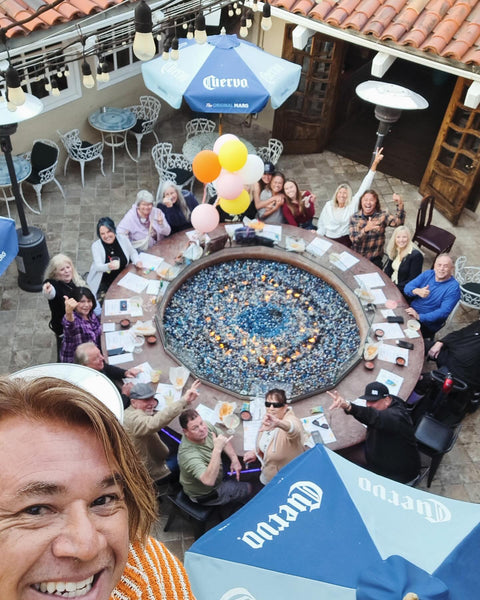 Guests gather with drinks around a circular fire pit on La Capilla's patio, beneath blue umbrellas and string lights, with colorful balloons.