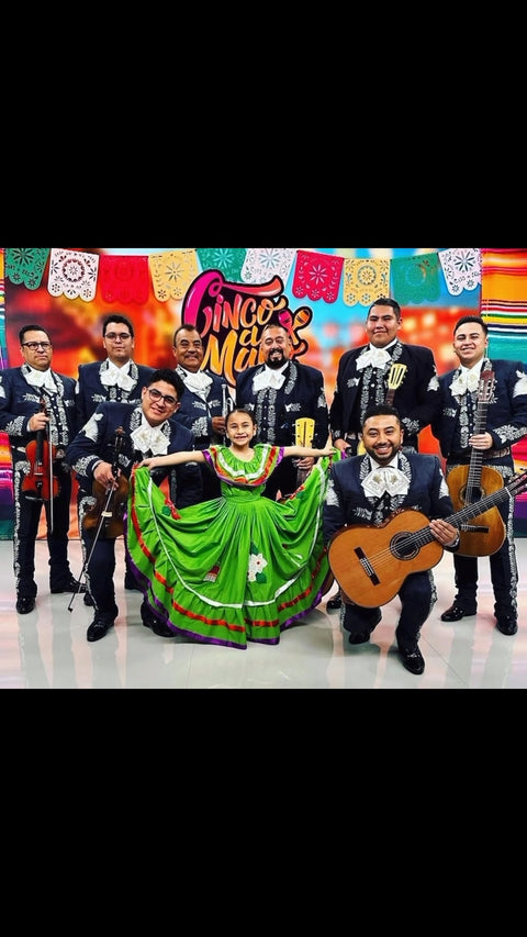 Mariachi musicians pose with a dancer in a bright green dress beneath colorful papel picado and a "Cinco de Mayo" banner.
