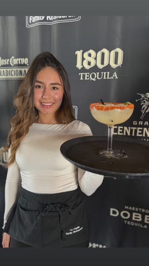 A server holds a tray with a margarita garnished with a red rim and a slice of lime, in front of a tequila-brand backdrop.